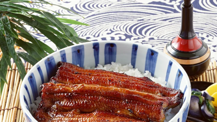 一碗白米饭上铺着色泽浓郁的蒲烧鳗鱼，旁边摆有绿叶和传统器具。