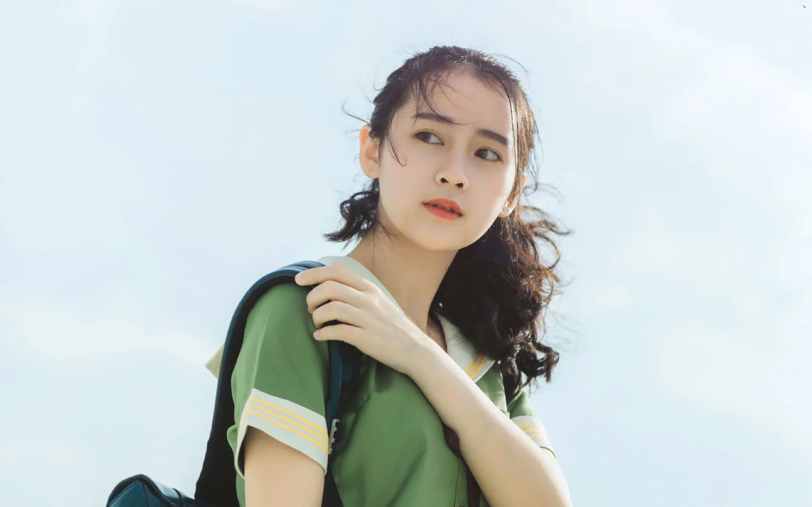 一名身穿绿色上衣、背着背包的女性站在明亮的蓝天下。