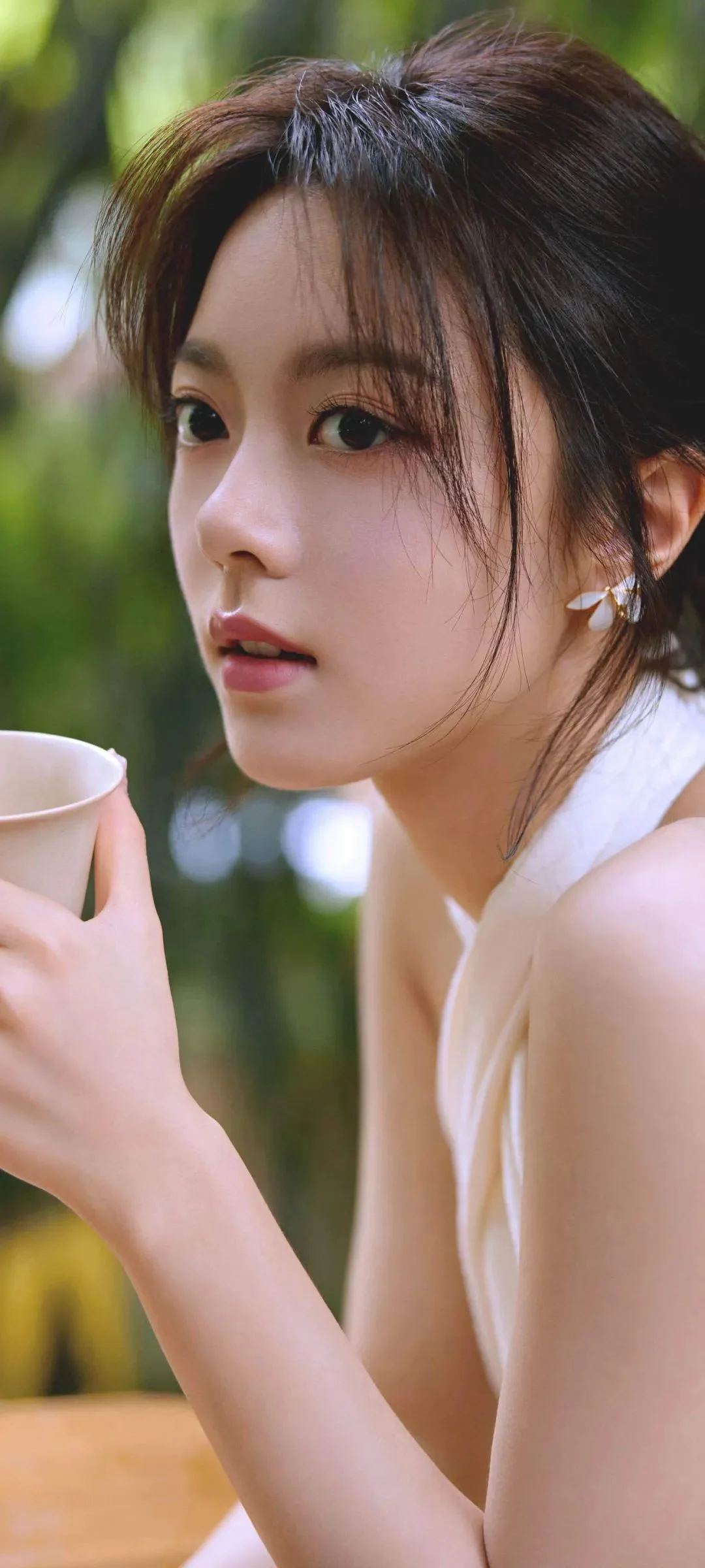 한 여성이 야외에서 컵을 들고 있으며 배경에는 흐릿한 녹색 식물이 보입니다.
