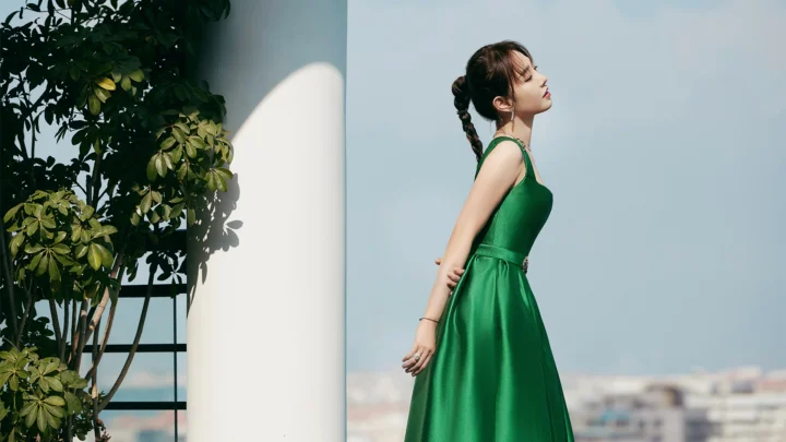 초록색 드레스를 입은 여성이 흰색 기둥 옆에 서 있고 배경에는 식물과 푸른 하늘, 멀리 건물이 보인다.