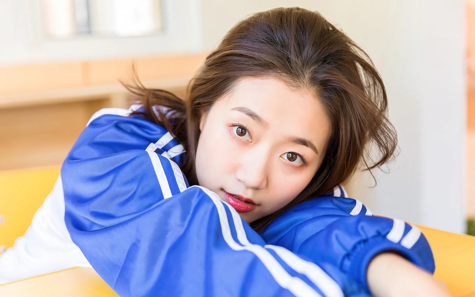 一名女性穿着蓝色带白色条纹的运动外套，在室内俯身看向镜头。