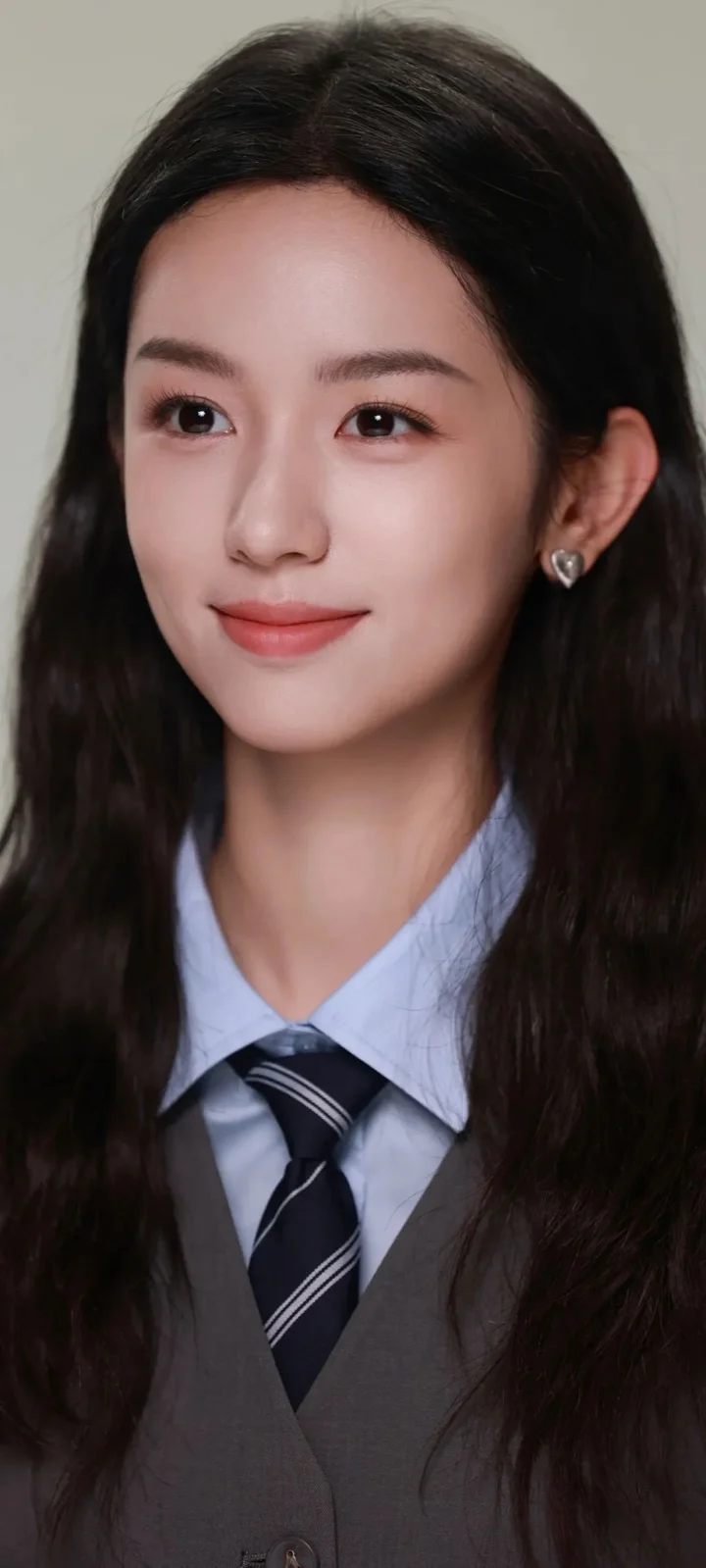 一名长发女性身穿浅蓝色领衬衫、深色领带和灰色背心，对着镜头微笑。