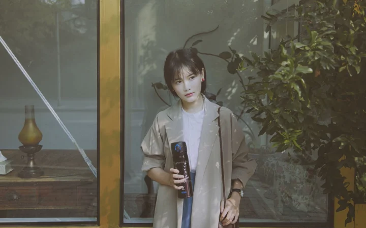 一名女子站在窗边，手持物品，身旁可见绿植。