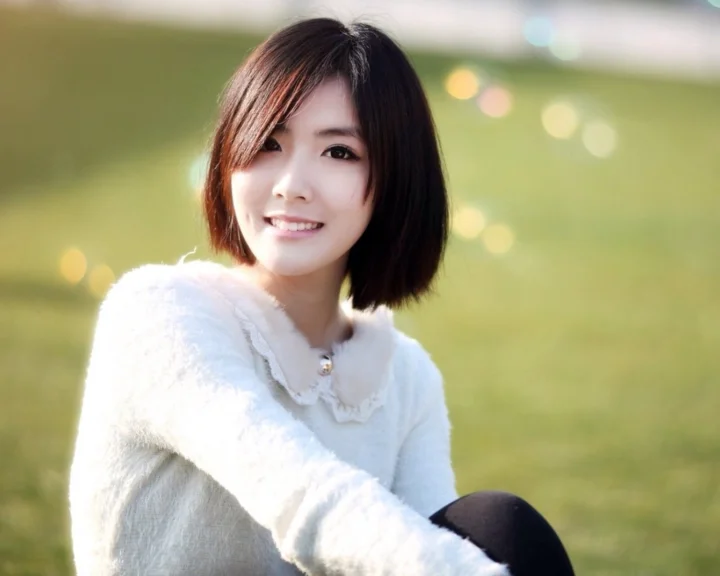 一位短发女子穿着白色毛衣坐在阳光明媚的草地上微笑。