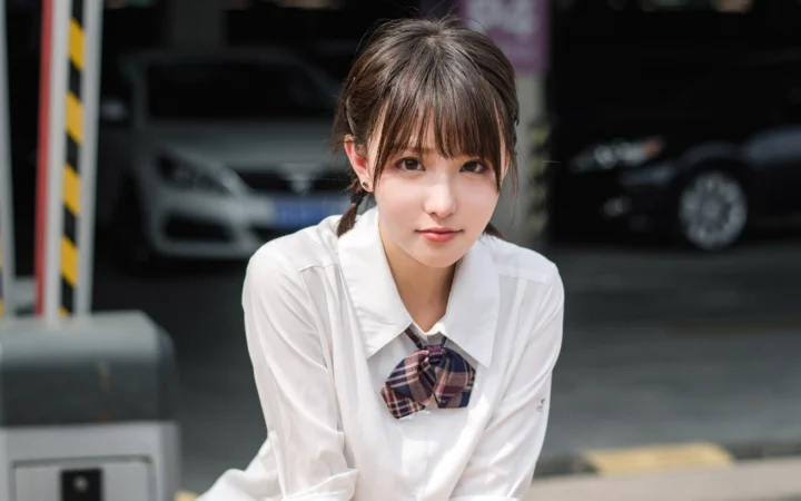 흰 셔츠와 체크무늬 나비넥타이를 착용한 성인 여성이 차량이 주차된 실내 공간 앞에 서 있습니다.