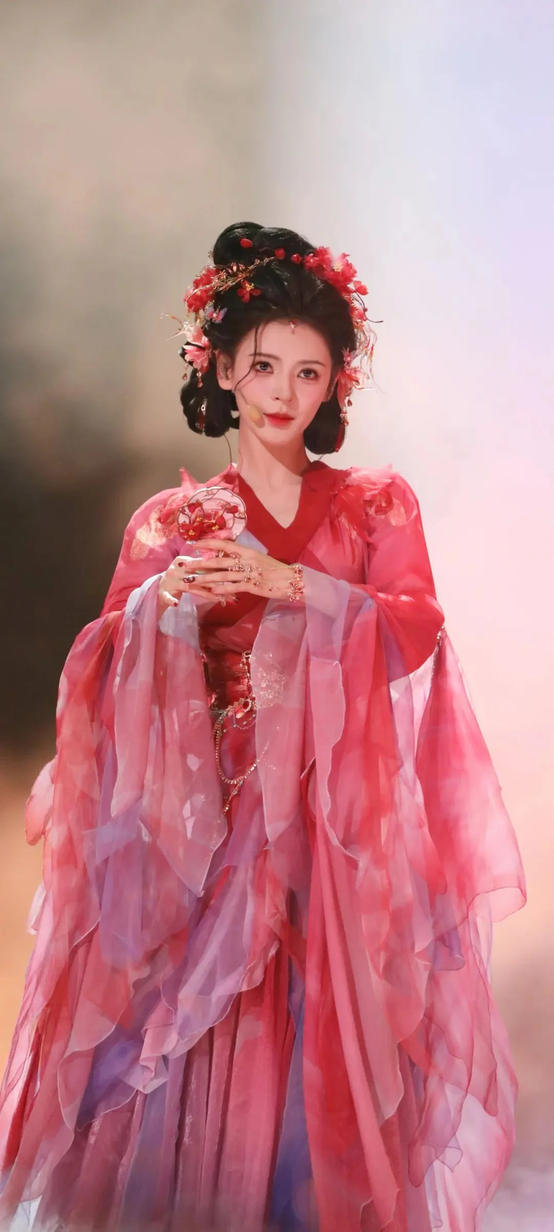 붉은 전통 의상과 꽃 장식을 착용한 여성이 부드러운 분홍색 배경 앞에 서 있습니다.