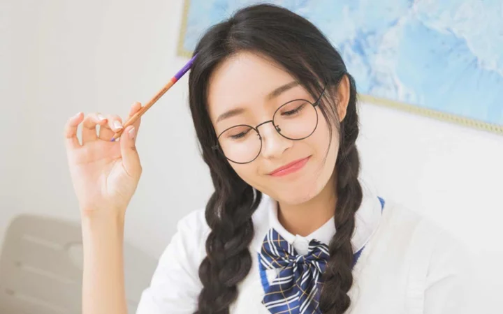 둥근 안경과 양갈래 땋은 머리를 한 젊은 여성이 연필을 들고 카메라를 향해 미소 짓고 있습니다.