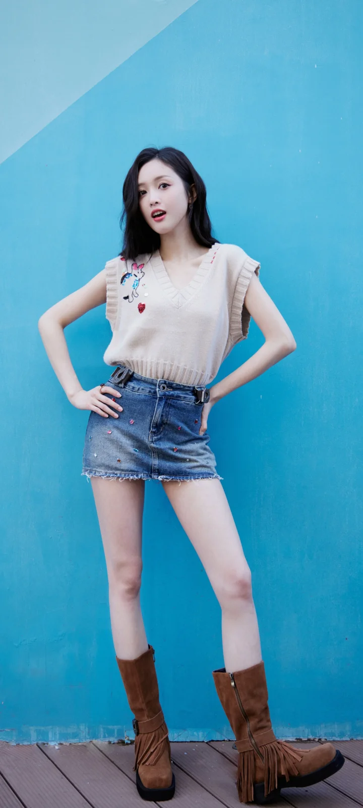 一名成年女性站在蓝色墙面前，穿着无袖上衣、牛仔短裙和棕色长靴。