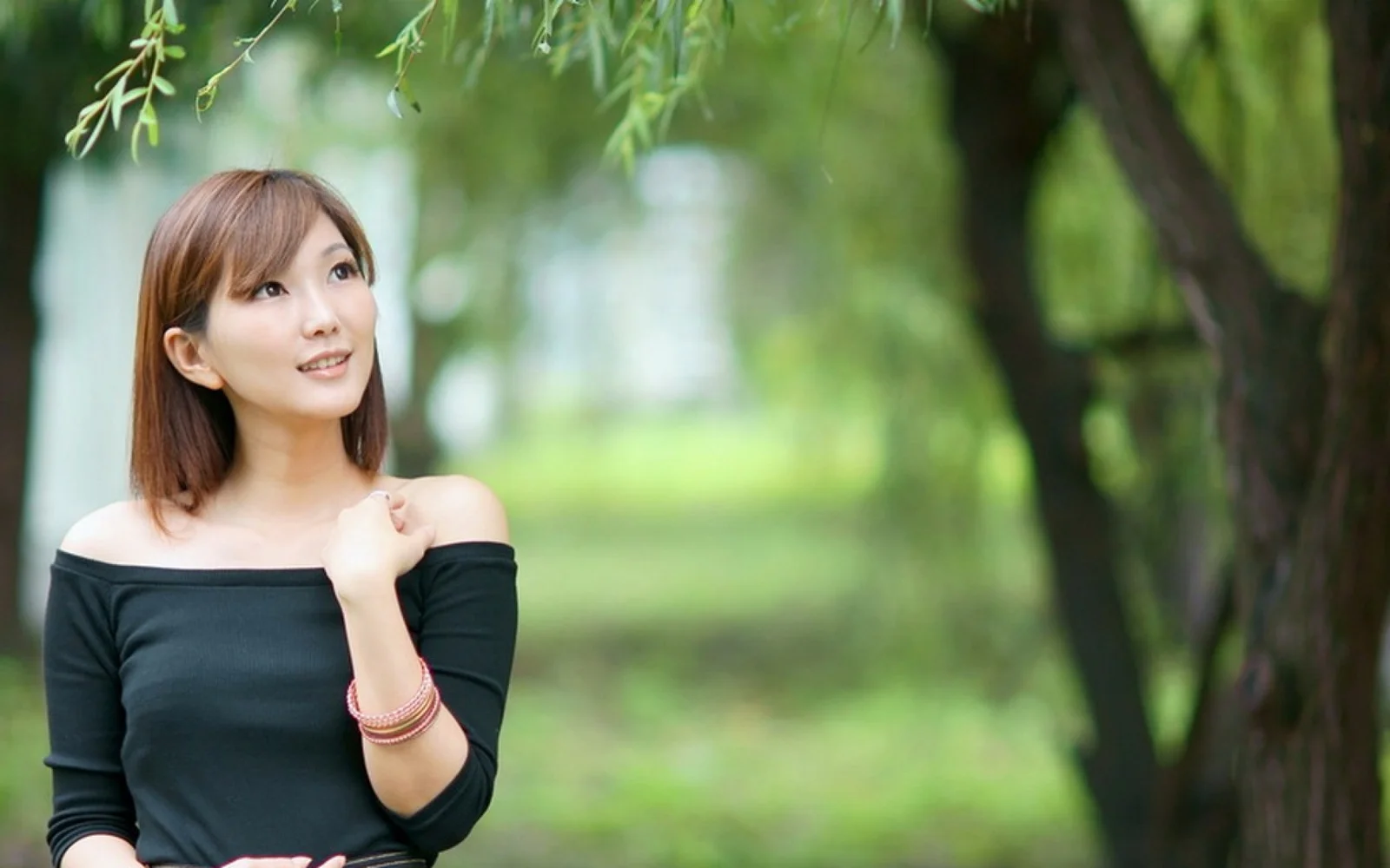一名棕色短发女性穿着黑色露肩上衣站在绿意盎然的树木之间。