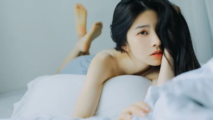 一名黑发女性俯卧在白色床品上，面向镜头。