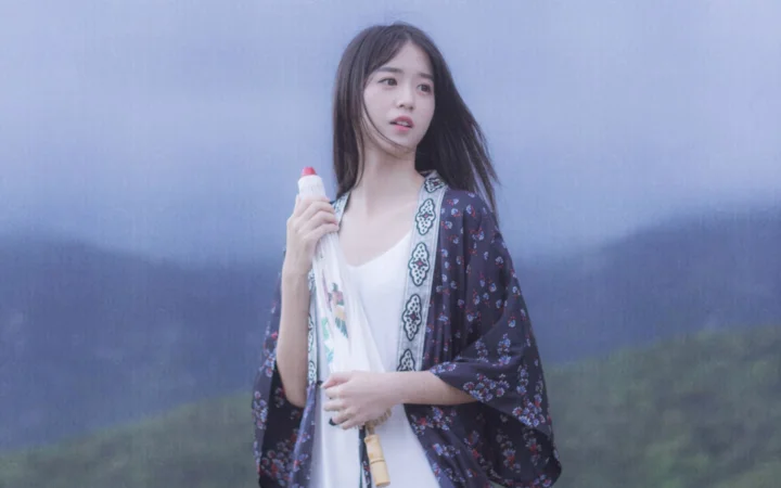 一名女子站在朦胧的山景前，手持一把白色雨伞。