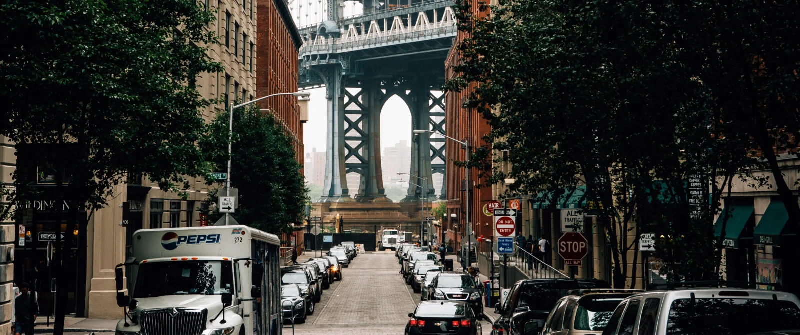 一条两侧停满汽车的城市街道通向远处的大型钢结构桥梁，周围是建筑和树木。