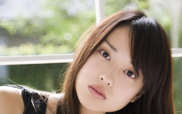 茶色の長い髪の女性が窓辺に寄り添い、外には柔らかな緑の景色が見えます。