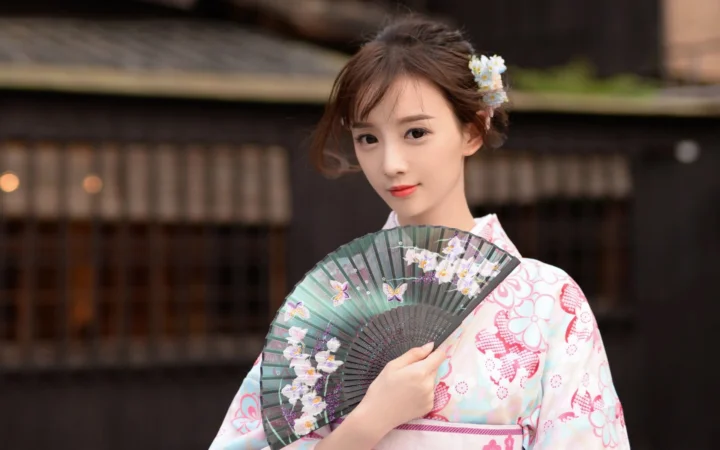 一位成年女子身穿浅色和服，手持绘有花朵与蝴蝶图案的折扇，站在传统建筑前。