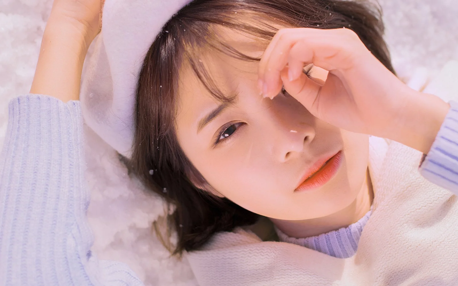 연보라색 니트를 입은 여성이 하얗고 푹신한 표면에 누워 얼굴 가까이에 팔을 둔 채 카메라를 바라보고 있다.