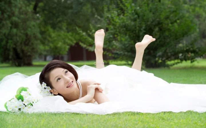 一名身穿白裙的女子躺在草地上，身旁放着一束白色小花，背景是绿色树林。