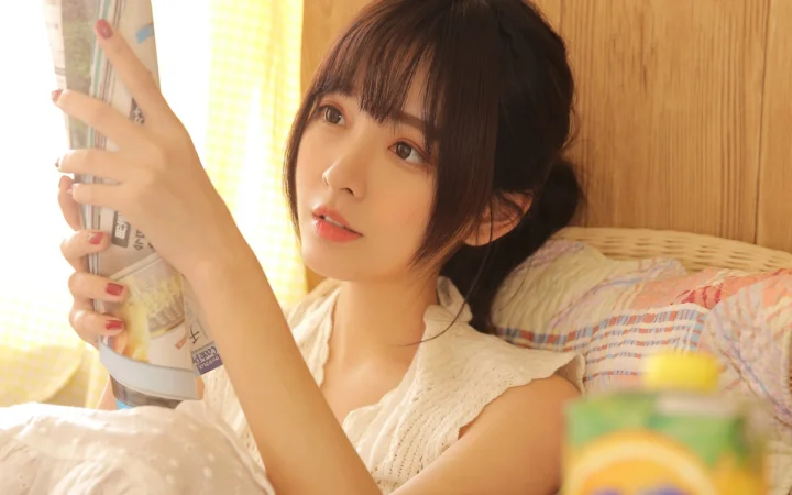 一位成年女性坐在室内窗边，手持杂志看向镜头。