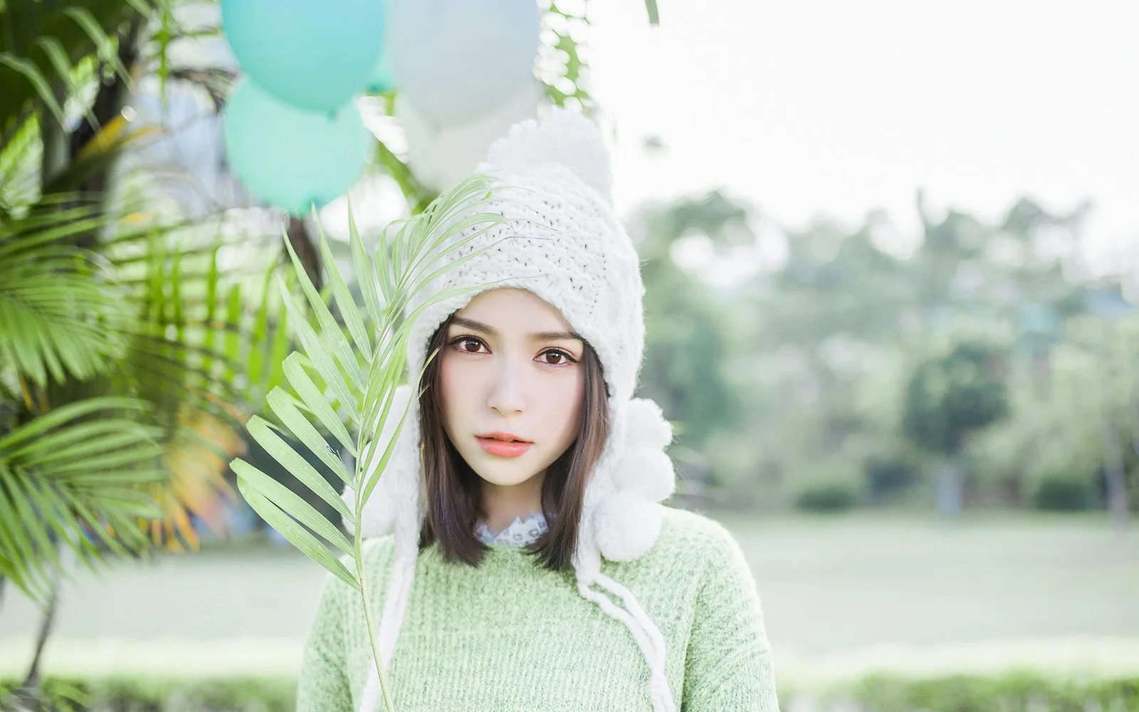 一名戴白色针织帽的女性站在户外绿植与湖景前，手边有一片棕榈叶。