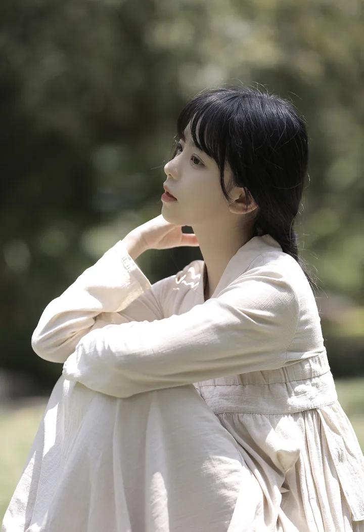 一名身穿白色连衣裙的女性坐在户外树影中侧头凝望。