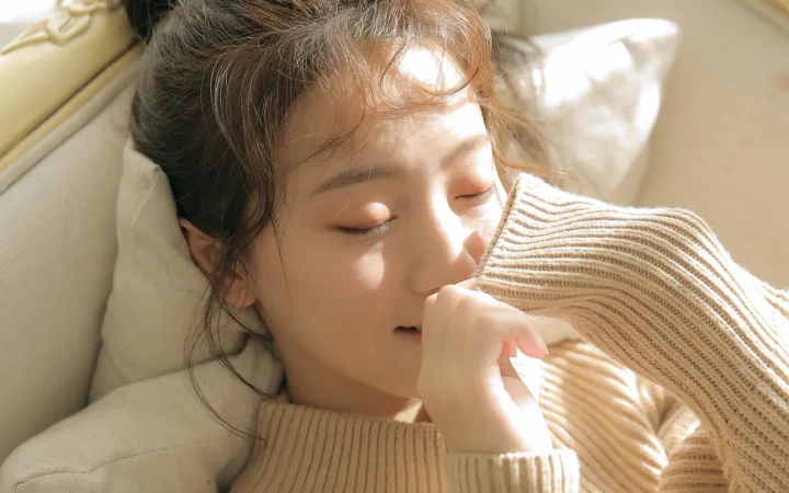 一名穿着针织毛衣的女子闭眼靠在枕头上，沐浴在温暖的阳光中。