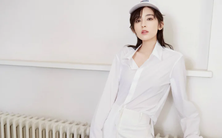 一位戴帽子的成年女性身穿白衬衫站在室内浅色墙壁前。