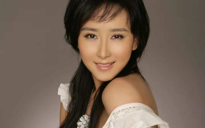 一位短发女性身穿白色露肩上衣，在灰色背景前微笑面对镜头。