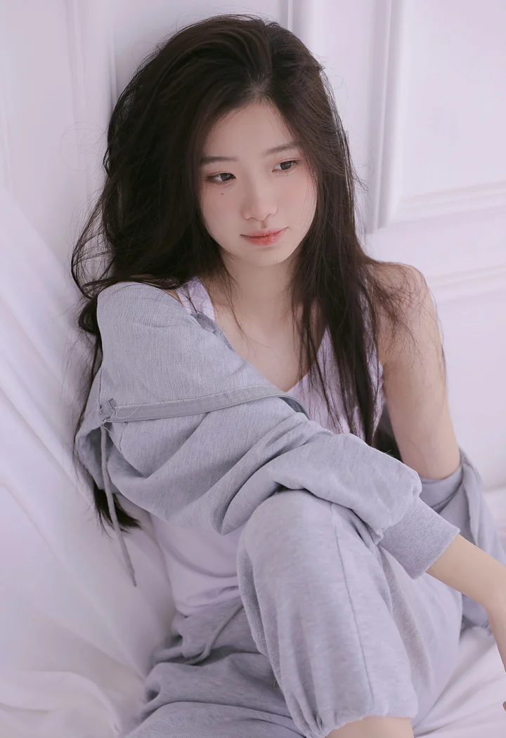 一名长发女性穿着宽松的灰色服装，坐在柔和淡紫色调的室内床上。