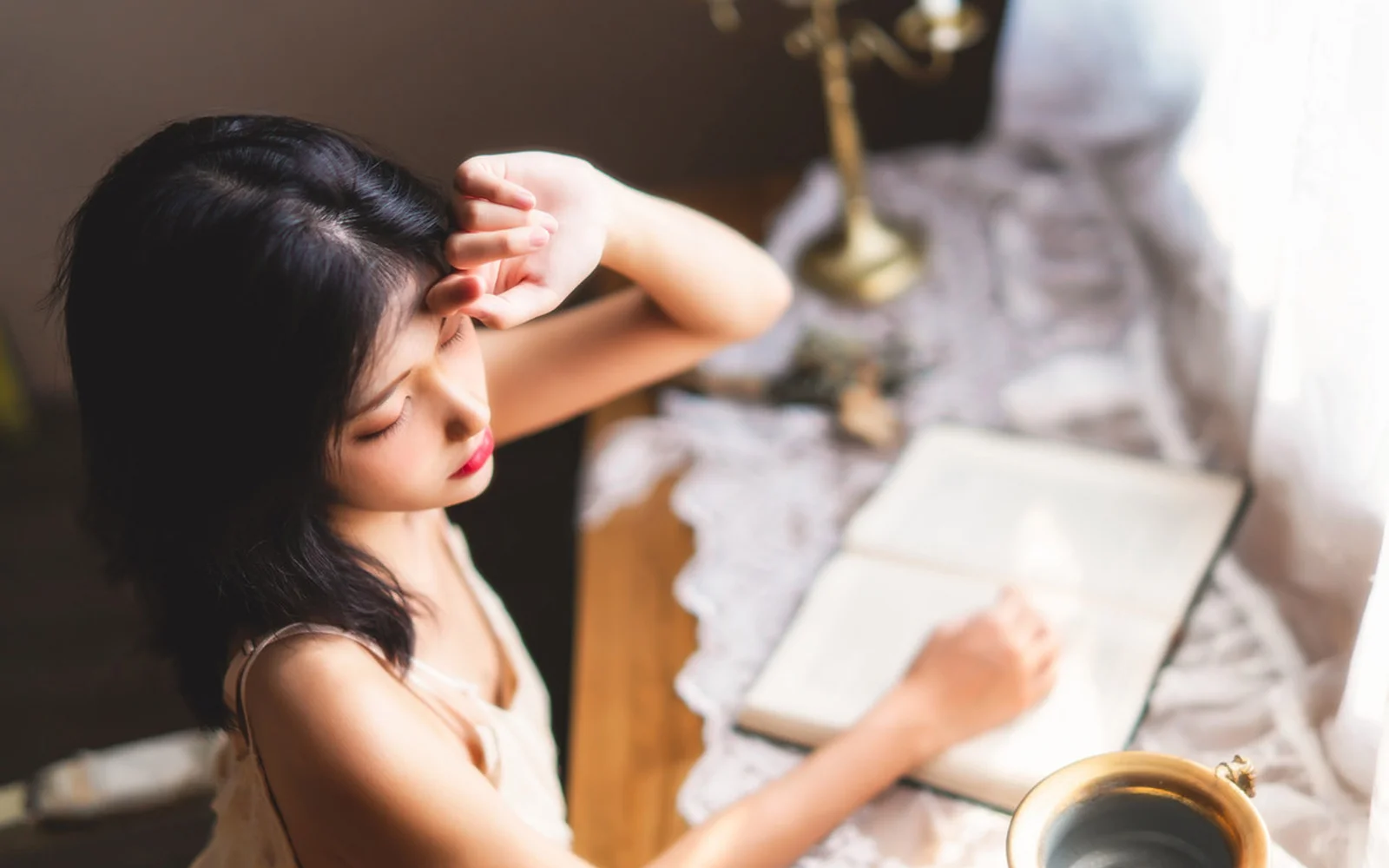 한 여성이 테이블 앞에 앉아 펼쳐진 노트와 커피잔 곁에서 한쪽에서 들어오는 밝은 빛을 받고 있습니다.
