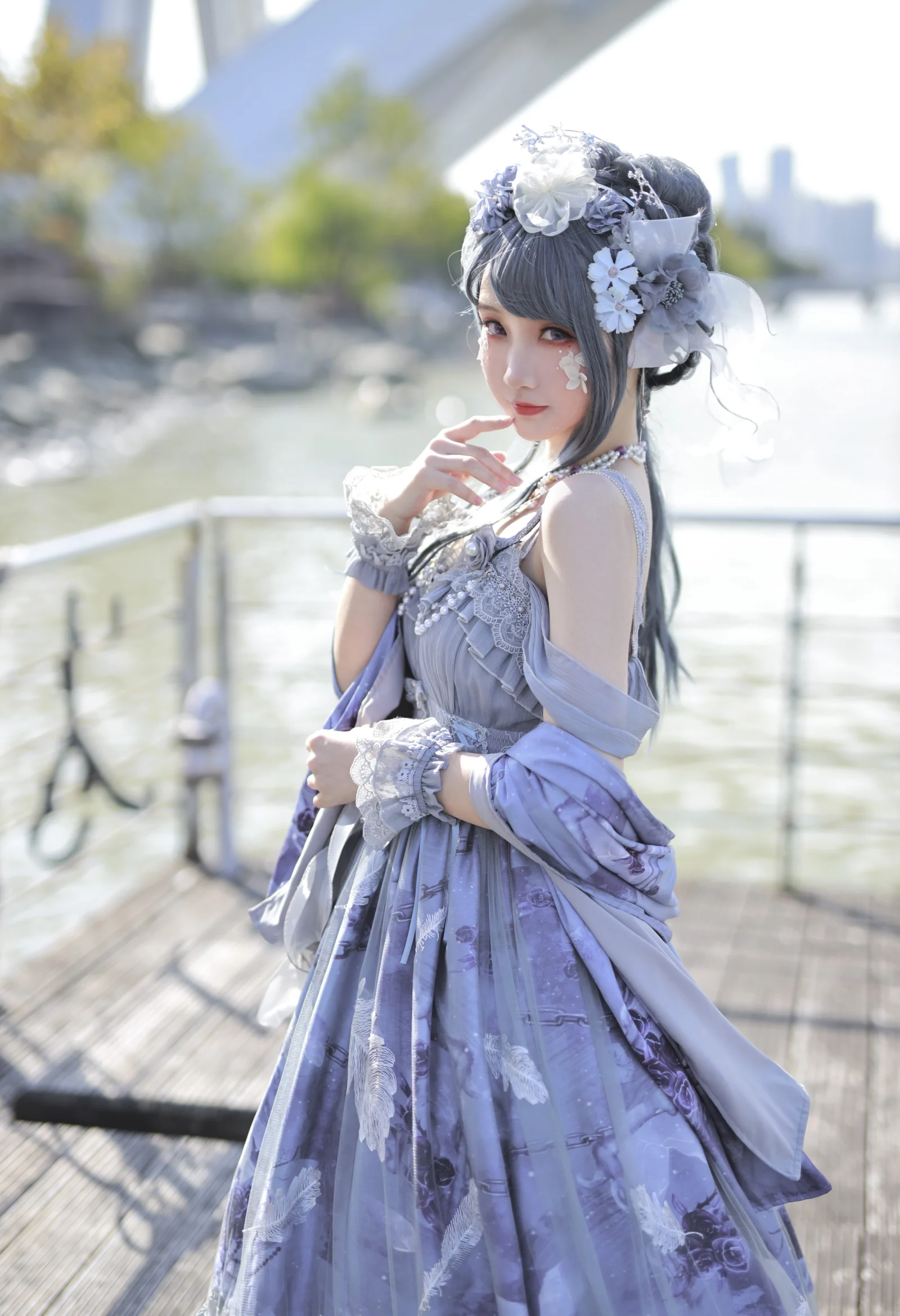 花飾りをつけてラベンダー色のドレスを着た女性が、水辺の遊歩道に立っている。