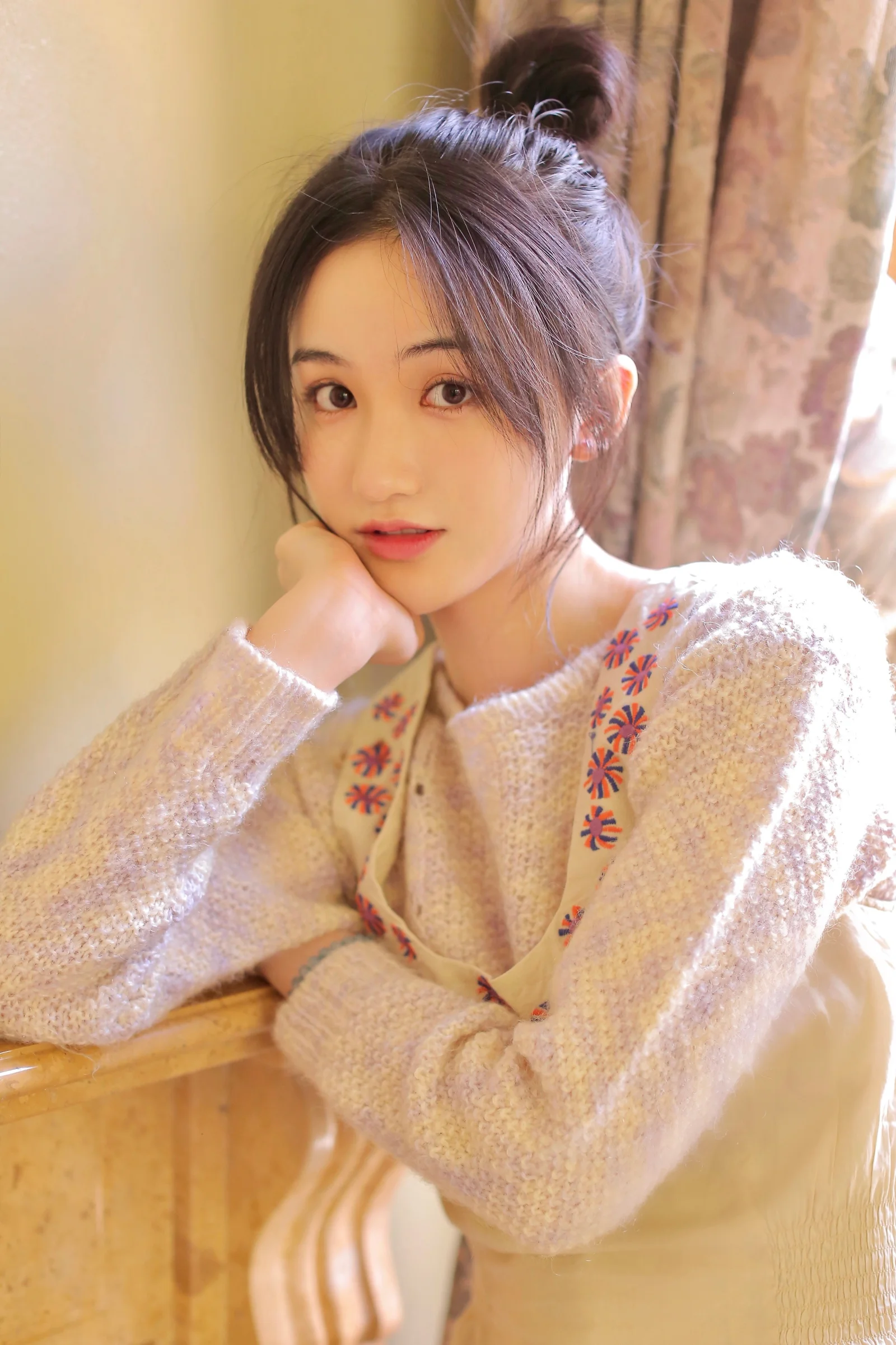 一位穿着浅色针织衫的女性靠在窗边，以手托腮沐浴在柔和阳光中。