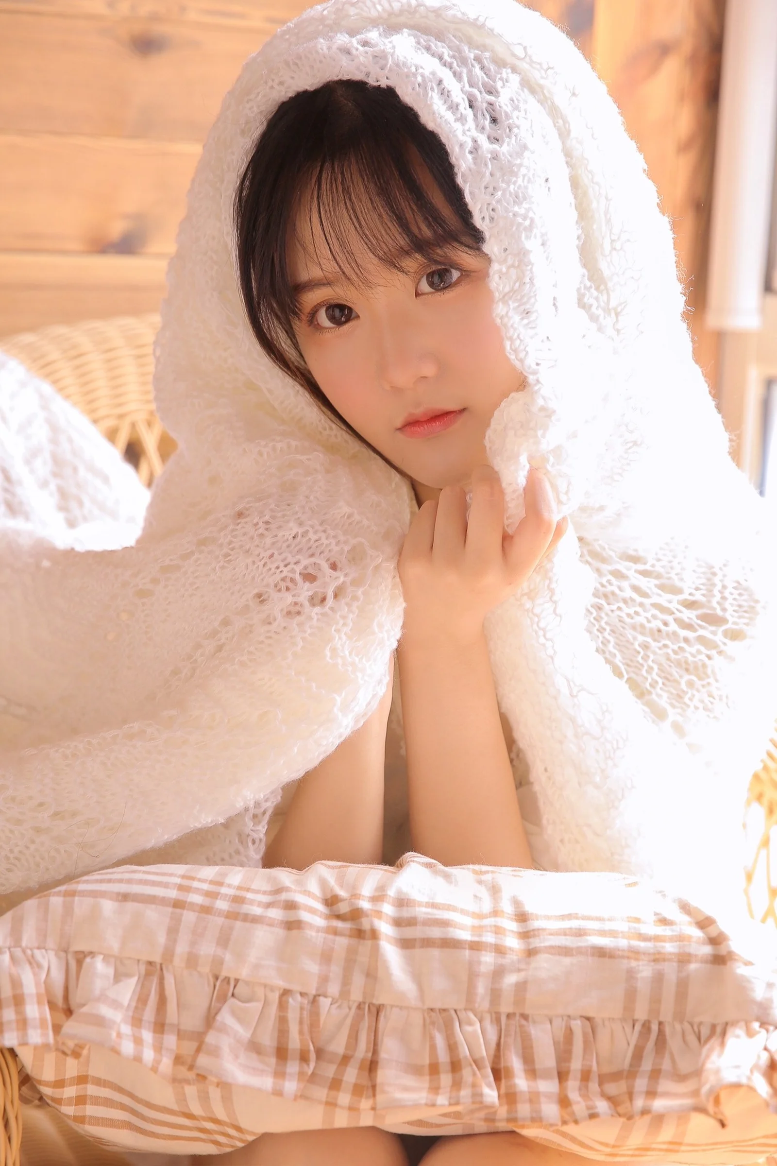 白いニットのブランケットに包まれた女性が、室内でチェック柄の寝具を前景にして座っている。