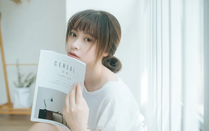 밝은 창가에서 여성이 잡지를 들고 카메라를 바라보고 있습니다.