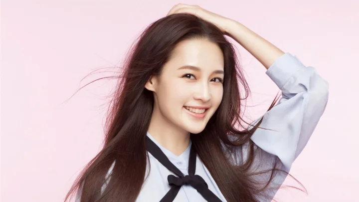 一名长发女子在浅粉色背景前微笑并抬手整理头发。