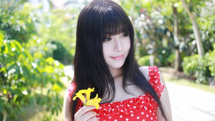 一名黑色长发女子身穿红色波点连衣裙，在阳光明媚的花园中手持黄色花朵。
