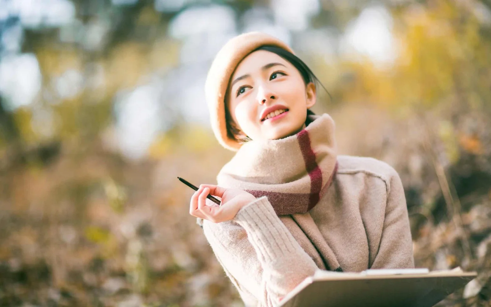 一位戴着围巾的女性在秋日户外手持笔记本微笑。
