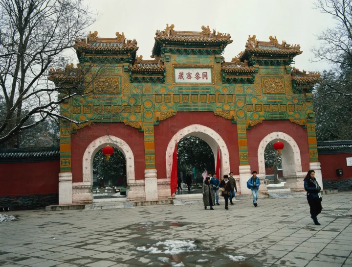 一座色彩鲜艳的中式古建筑牌楼矗立在铺石庭院中，周围可见树木和少量积雪。