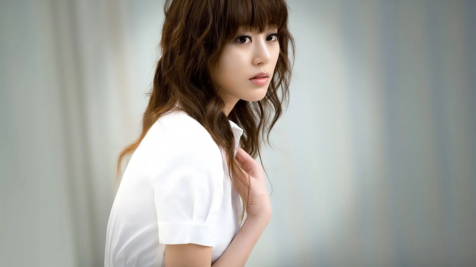 흰 셔츠를 입은 성인 여성이 부드러운 실내 배경 앞에서 옆으로 서서 카메라를 바라보고 있습니다.