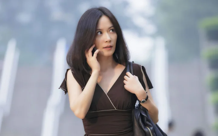 어두운 원피스를 입은 여성이 흐릿한 도시 배경 앞에서 검은색 핸드백을 들고 서 있습니다.