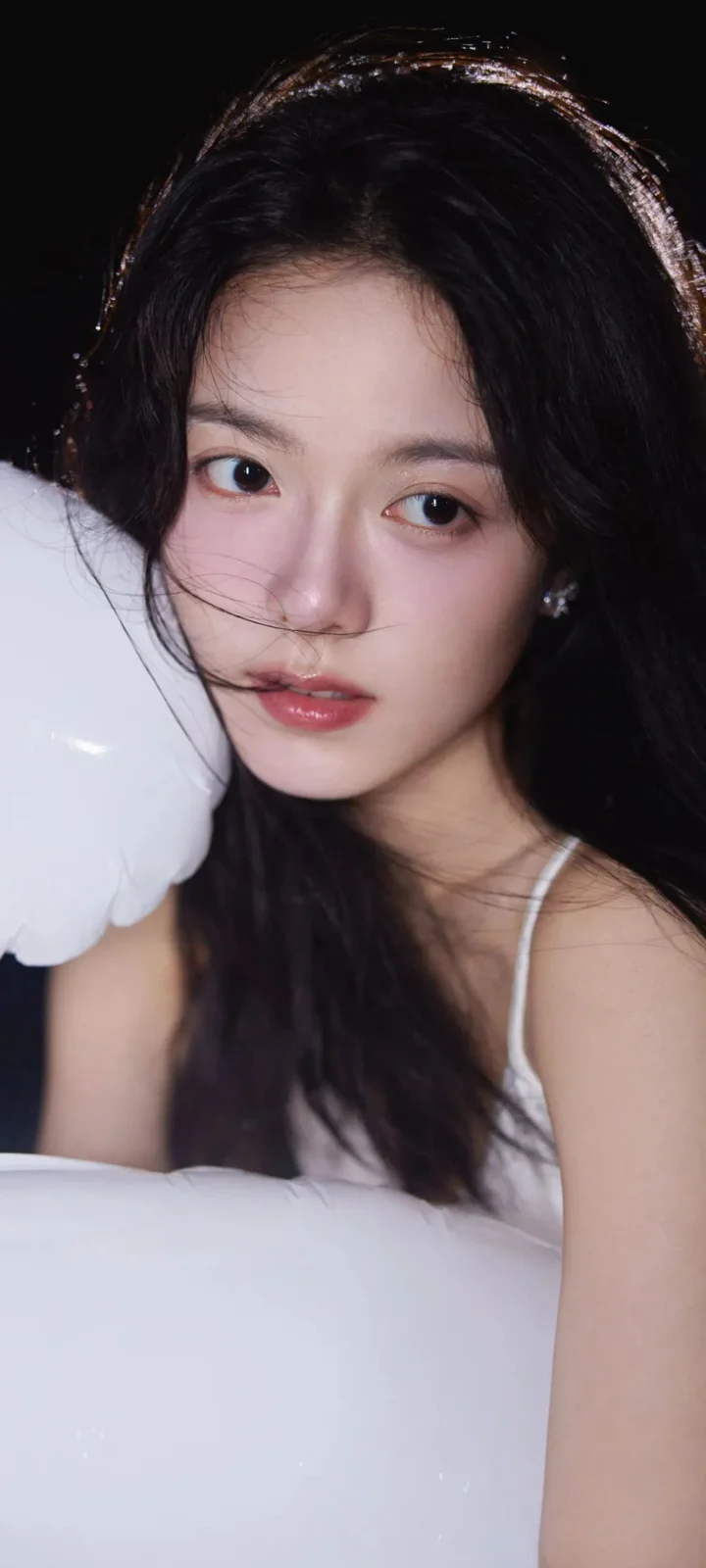 一名长发女性靠在白色枕头旁，身穿浅色细肩带上衣，黑色背景衬托出她的面部与上半身。