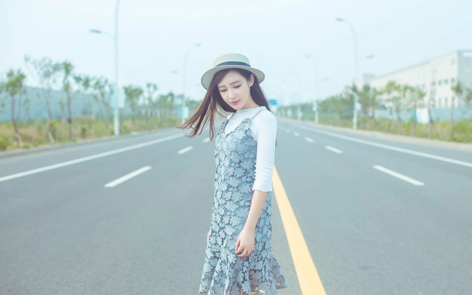 一名戴着帽子的女子站在宽阔道路中央，背景是淡蓝色天空和道路两侧的树木与建筑。