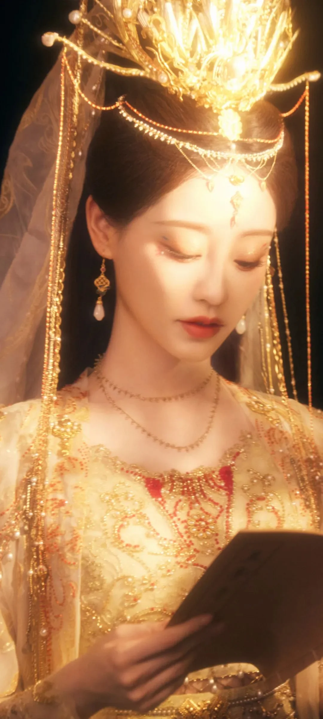 一名成年女性身穿华丽的金色传统服饰，佩戴繁复头饰和珠宝，低头手持深色物件。