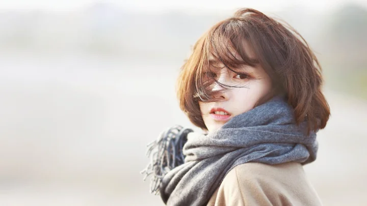 一位短发女性裹着灰色围巾回望镜头，背景明亮而虚化。