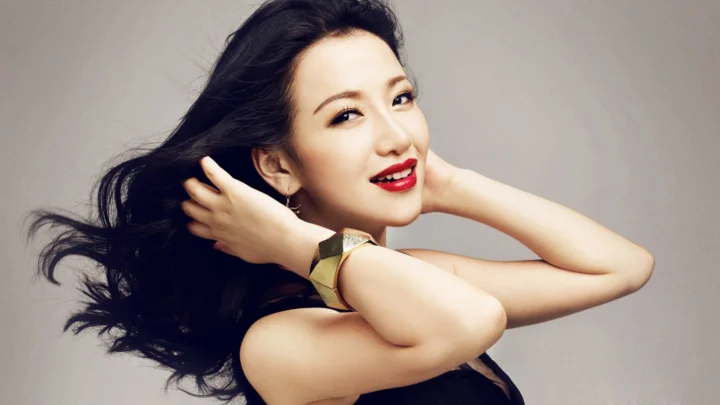 一位黑发女性在灰色背景前整理头发并面向镜头微笑。