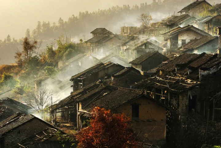 层叠的传统民居沿山坡铺展开来，晨雾、树木与暖色光线交织成宁静的乡村景象。