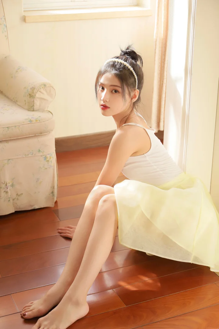 一名扎着发髻并戴着发饰的女子穿着白裙坐在明亮的室内木地板上。