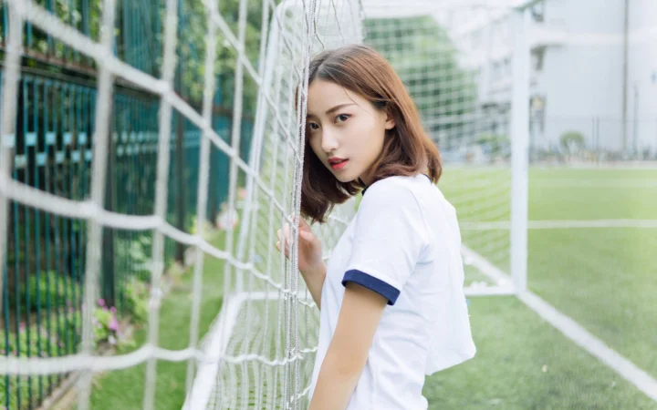 一名短发女子站在足球场旁的球网边，身后是绿色场地和建筑。