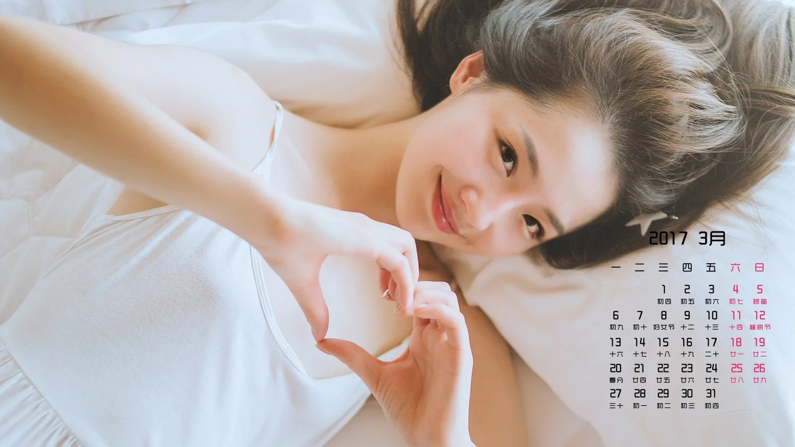 성인 여성이 흰색 침대에 누워 양손으로 하트 모양을 만들고 있다.