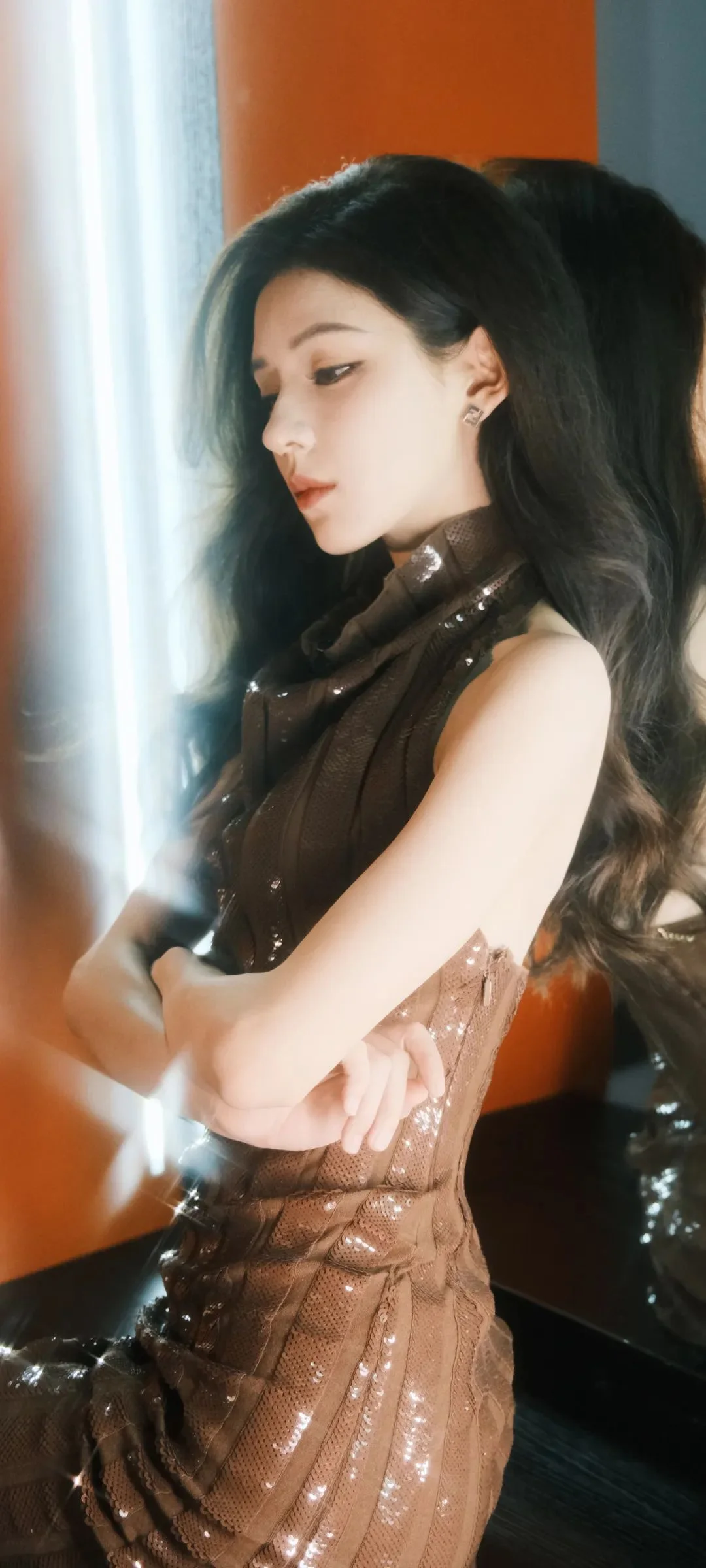 一名长发女性身穿棕色亮片礼服侧身站在室内窗边。