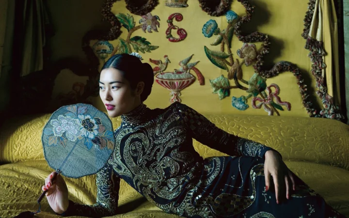 一名成年女性身穿深色刺绣服饰，手持折扇斜倚在沙发上。