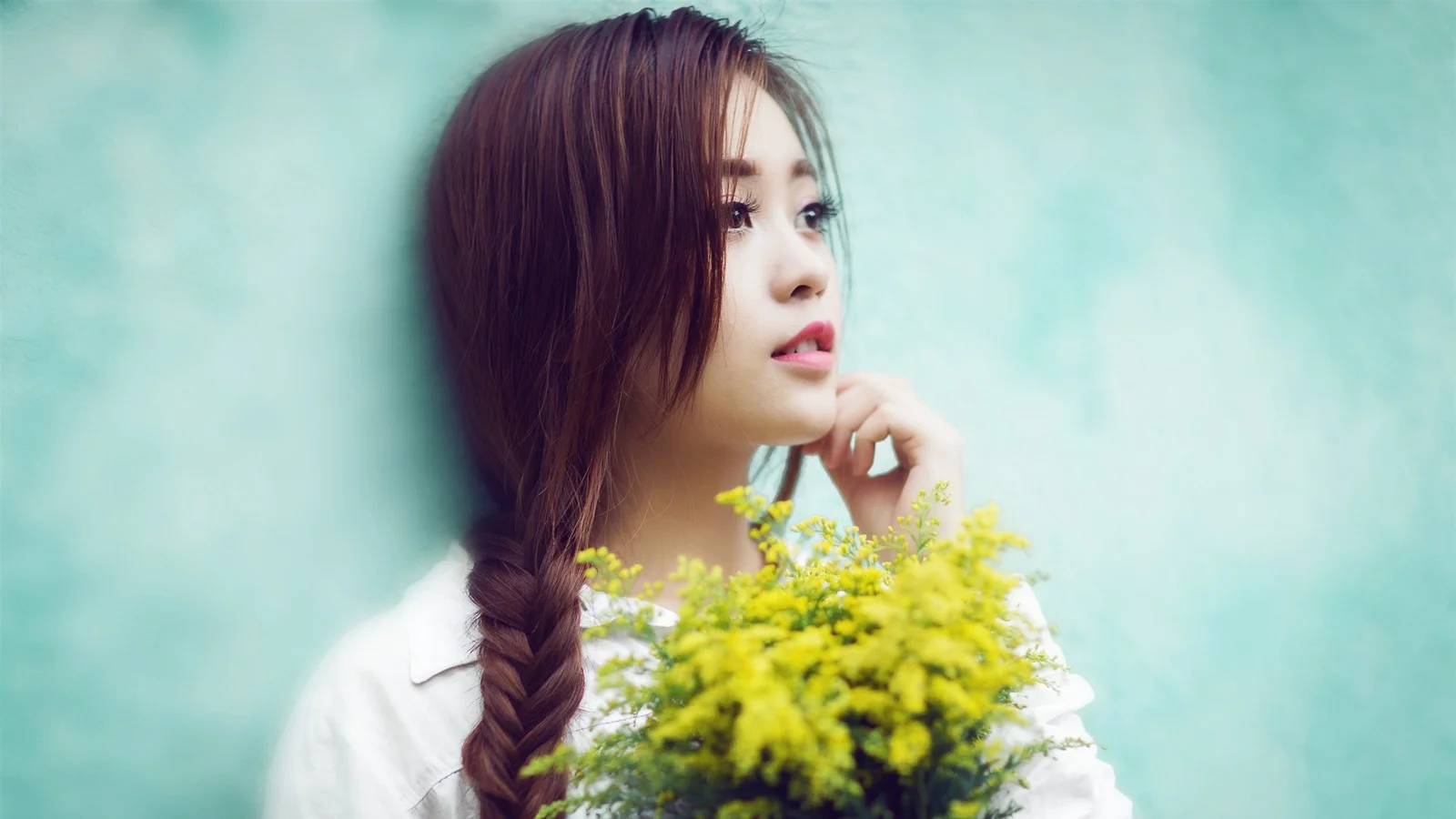 긴 땋은 머리의 여성이 노란 꽃다발을 들고 부드러운 청록색 배경을 바라보고 있습니다.